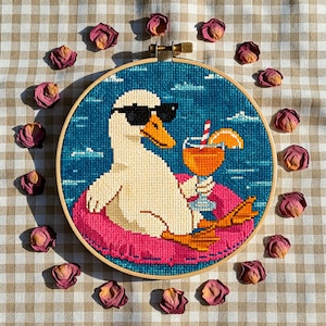 May include: A circular embroidery hoop containing a cross-stitch pattern of a white duck wearing black sunglasses, floating on a bright pink inner tube in blue water, holding an orange cocktail with a straw and a slice of fruit garnish.