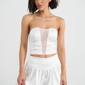 May include: A white two-piece set consisting of a strapless satin crop top with a vertical sheer mesh panel at the center and a matching high-waisted mini skirt featuring a gathered, voluminous bubble-style hemline and a smooth waistband.