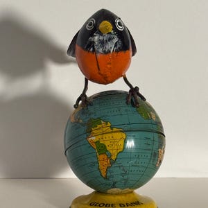 May include: A metal coin bank featuring a robin bird with a black head, white throat, and orange chest perched atop a spherical globe. The globe displays a map of South America and the Atlantic Ocean, with the text GLOBE BANK printed on the yellow base.