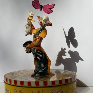 May include: A clown figurine standing on a round yellow tin with carousel horse illustrations. The clown wears an orange coat, striped trousers, and a green tie, holding a white rabbit in one hand and a black top hat in the other, with a pink butterfly.