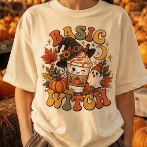 Può includere: Una t-shirt color avorio con una stampa grafica che riporta il testo BASIC WITCH. Il design include una tazza di caffè con un cappello da strega, una zucca, un piccolo fantasma, foglie autunnali e una luna crescente nei toni dell'arancione e giallo.