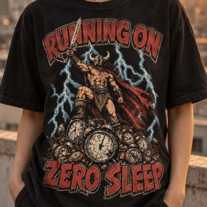 May include: A black short-sleeve t-shirt featuring a graphic print of a warrior in a horned helmet holding a sword, standing atop a pile of clocks. The text "RUNNING ON" appears above and "ZERO SLEEP" below in red, with blue lightning bolts.
