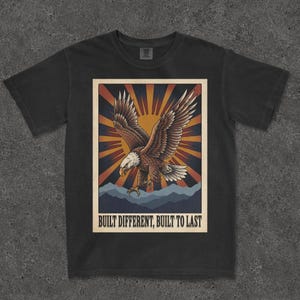 Può includere: Una t-shirt nera a maniche corte con una stampa grafica di un'aquila calva in volo sopra sagome di montagne. Lo sfondo mostra un motivo a raggiera in arancione e giallo. Il testo BUILT DIFFERENT, BUILT TO LAST è stampato in basso.
