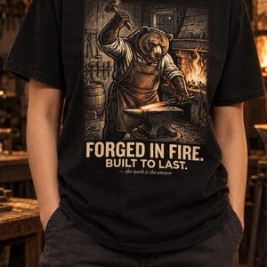 Puede incluir: Una camiseta negra con una ilustración gráfica de un oso con delantal trabajando en un yunque dentro de una herrería. El texto dice: FORGED IN FIRE. BUILT TO LAST. — the work is the answer. El diseño incluye una fragua encendida.