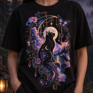 May include: A black t-shirt featuring a graphic print of a black cat sitting before a crescent moon and a full moon. The design includes purple and blue clouds, flowers, butterflies, hanging ornaments, and a mountain landscape with a waterfall.
