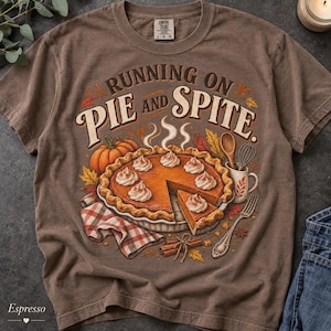 Op de afbeelding: Een bruin t-shirt met korte mouwen en een afbeelding van pompoentaart met slagroom, een pompoen, een garde, een lepel en kaneelstokjes. De tekst luidt RUNNING ON PIE AND SPITE. Het shirt heeft een ronde hals en een Comfort Colors label.