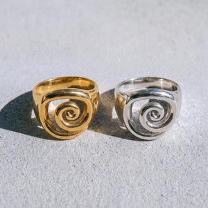 May include: Two rings, one gold-toned and one silver-toned, each featuring a prominent central spiral design. The rings have a smooth, polished finish and a thick, rounded band structure. The word abyss. is printed in black at the bottom center.