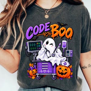 Puede incluir: Una camiseta gris carbón con un gráfico de un fantasma blanco que lleva un estetoscopio y hace el signo de la paz. El diseño incluye un carro médico morado, un monitor cardíaco, una bolsa de suero y dos calabazas con el texto CODE BOO.