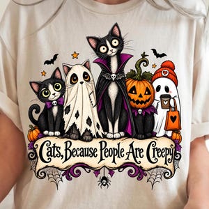 May include: A beige t-shirt featuring a graphic of four cats in Halloween costumes, including a ghost, a vampire, and a pumpkin-headed cat. The text "Cats, Because People Are Creepy" is printed on a scroll banner below the cats with spider web accents.