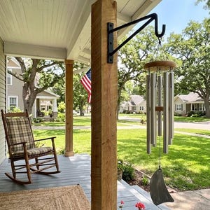 May include: A set of silver-toned metal wind chimes with a circular wooden top and a curved metal wind catcher, suspended from a black metal hook attached to a wooden porch post. The chimes feature multiple cylindrical tubes of varying lengths.