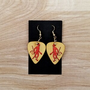 May include: A pair of wooden guitar pick earrings with a red devil design. The earrings are gold in color and have a teardrop shape.