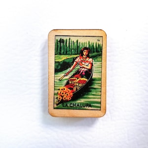 May include: A wooden card with a colorful illustration of a woman in a boat with flowers and fruit. The card reads "La Chalupa" and has a trademark symbol.