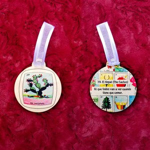 May include: Two round wooden ornaments with a white background and colorful illustrations. The ornament on the left features a green cactus with red flowers and the text "39 El Nopal". The ornament on the right features a colorful illustration of a cactus, a horseshoe, a bucket, and a pine tree. The text reads "39. El Nopal (The Cactus) Al que todos van a ver cuando tiene que comer."