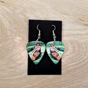 May include: A pair of guitar pick earrings with a colorful image of a woman in a boat on a lake. The earrings are made of wood and have gold-colored hooks.