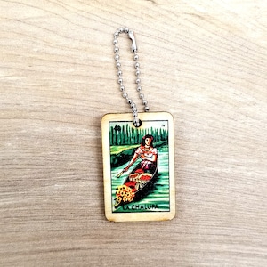 May include: A wooden keychain featuring a colorful illustration of a woman in a boat with flowers, with the text "La Chalupa" at the bottom. The keychain is attached to a silver chain.
