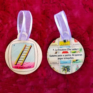 May include: Two round wooden ornaments with a colorful design. The ornament on the left features a yellow ladder with a pink background. The ornament on the right features a colorful game board with the text "7. La Escalera (The ladder) Subeme paso a pasito. No quieras pegar brinquitos."