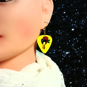 May include: A pair of earrings featuring a yellow guitar pick with a red, black, and green illustration of a woman wearing a hat. The earrings are gold.
