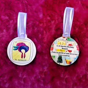 May include: Two round, white, wooden ornaments with a purple ribbon. The ornament on the left has a yellow background with a red and white hat on it. The text "13" and "EL GORRITO" are printed on the ornament. The ornament on the right has a colorful background with a picture of a tree and a bucket. The text "13. El Gorrito (The Baby Bonnet)" and "Ponle su gorrito al nene no se nos vaya a resfriar." are printed on the ornament.