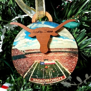 May include: A round wooden Christmas ornament with a picture of a football stadium and a Longhorns logo. The ornament is decorated with the text "Austin Texas".