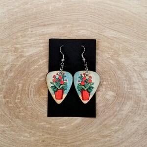 May include: A pair of earrings featuring a red flower design in a pot on a guitar pick shaped piece of wood.