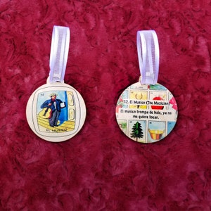 May include: Two round wooden ornaments with a colorful design. The ornament on the left features a cartoon illustration of a musician playing a guitar. The ornament on the right features a colorful illustration of a musician playing a trumpet with the text "32. El Musico (The Musician) El musico trompa de hule, ya no me quiere tocar."