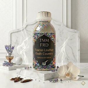 May include: A clear glass bottle of perfume oil with a metallic, iridescent neck and a black label featuring the text TMM FRD Tuscan Leather Oudh Country Perfume Oil 100 ml. The bottle is decorated with a colorful, intricate, floral-like pattern.