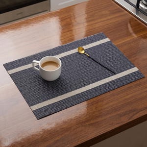 May include: A rectangular placemat featuring a dark blue and white herringbone pattern with two horizontal beige stripes. A white ceramic mug filled with a light brown beverage and a slender gold-toned spoon with a black handle rest on top.