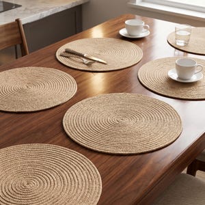 May include: A set of five round, tan-coloured woven placemats with a concentric spiral texture. The placemats are arranged on a wooden table, with some featuring white ceramic cups, saucers, a clear glass, and silver-toned cutlery placed on top.