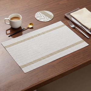 May include: A rectangular placemat and a round coaster featuring a white and gray herringbone pattern with two horizontal tan stripes. The set is accompanied by a white ceramic mug, a gold-toned spoon, a silver fork, a silver knife, and a folded napkin.