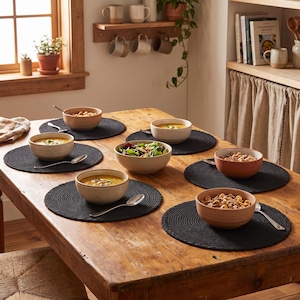 Pode incluir: Um conjunto de seis jogos americanos redondos de tecido preto dispostos sobre uma mesa de jantar de madeira rústica. Cada jogo americano contém uma tigela de cerâmica com diversos alimentos, incluindo sopa, salada e cereais, acompanhados por uma colher de metal prateado.