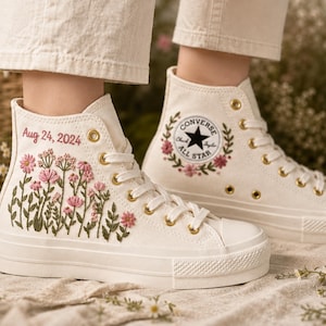 Könnte beinhalten: Cremefarbene High-Top-Sneaker aus Canvas mit gestickten rosa Wildblumen und grünen Stielen an der Seite. Der Text Aug 24, 2024 ist in Rosa gestickt. Die Schuhe haben goldene Ösen, weiße Schnürsenkel und das klassische Converse All Star Logo.