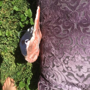 May include: A square purple pillow featuring a raised, velvet-like damask pattern in a darker shade of purple. Beside the pillow is a partially visible, light pink, textured object with a white cord, resting on a patch of green grass.