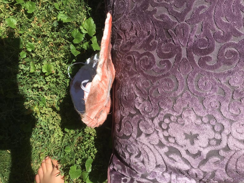 May include: A square purple pillow featuring a raised, velvet-like damask pattern in a darker shade of purple. Beside the pillow is a partially visible, light pink, textured object with a white cord, resting on a patch of green grass.