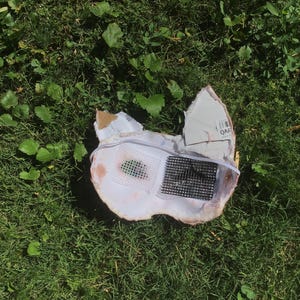 May include: A white, mask-like object with pointed ears, featuring a black grid-patterned mesh insert on the right side and a smaller, dark green mesh insert on the left side. The structure is made of a rigid material with a thin white elastic strap.