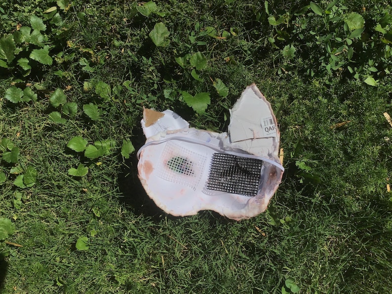 May include: A white, mask-like object with pointed ears, featuring a black grid-patterned mesh insert on the right side and a smaller, dark green mesh insert on the left side. The structure is made of a rigid material with a thin white elastic strap.