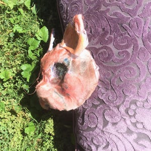 May include: A sculpted mask featuring a textured, flesh-toned surface with subtle pink and blue highlights. The mask includes pointed ears, a defined snout, and a dark, mesh-covered eye opening, presenting a detailed, three-dimensional animal-like form.