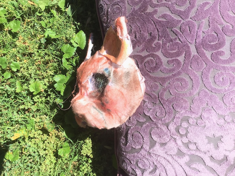 May include: A sculpted mask featuring a textured, flesh-toned surface with subtle pink and blue highlights. The mask includes pointed ears, a defined snout, and a dark, mesh-covered eye opening, presenting a detailed, three-dimensional animal-like form.
