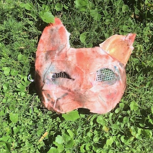 May include: A handcrafted animal mask featuring a textured, mottled surface in shades of orange, red, and pink. The mask has two pointed ears at the top and two eye openings covered with a fine, dark green grid mesh material for visibility.