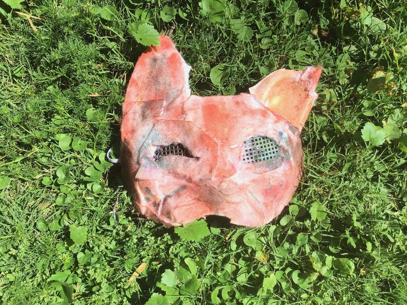 May include: A handcrafted animal mask featuring a textured, mottled surface in shades of orange, red, and pink. The mask has two pointed ears at the top and two eye openings covered with a fine, dark green grid mesh material for visibility.