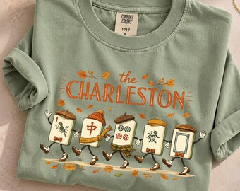 May include: A sage green short-sleeve t-shirt featuring a graphic of five anthropomorphic mahjong tiles wearing hats and scarves, walking among autumn leaves. The text reads the CHARLESTON in orange. A gold paw print pendant necklace is nearby.