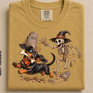 May include: A mustard-colored t-shirt featuring a graphic of a black and tan dachshund running with a bone in its mouth, pursued by a skeleton wearing a witch hat. The design includes two tombstones marked RIP and the text Comfort Colors size L.