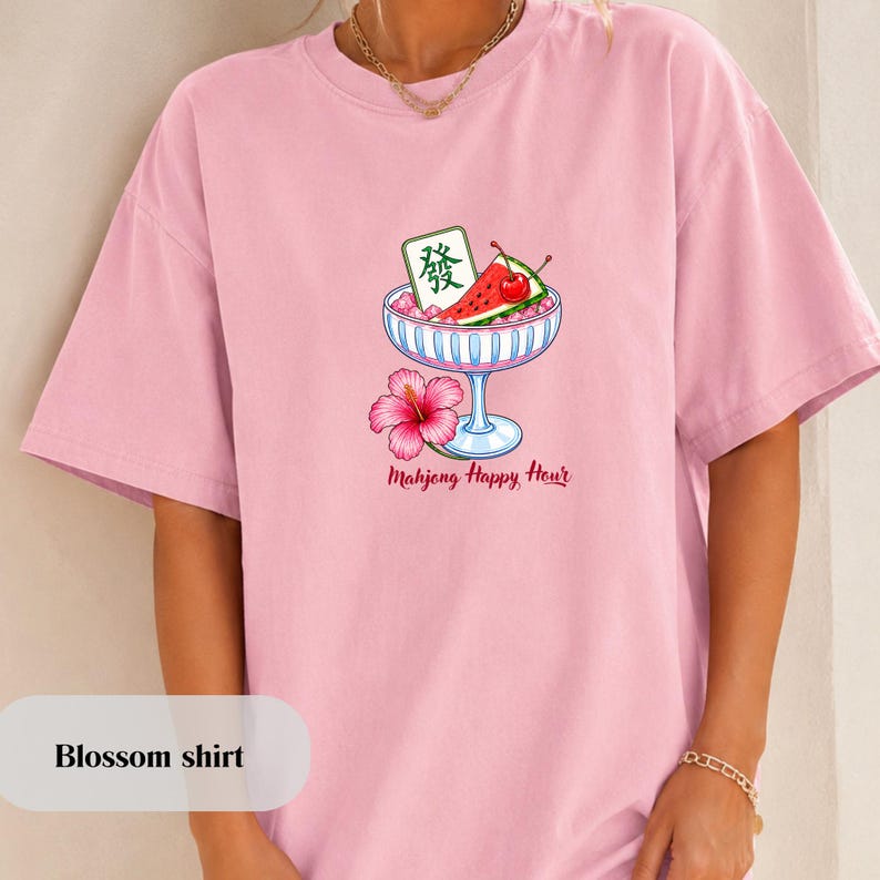 Peut inclure: Un t-shirt rose clair avec un motif repr&eacute;sentant une coupe en verre remplie de glace rose, une tranche de past&egrave;que, une cerise rouge et une tuile de mah-jong verte. Une fleur d'hibiscus rose est &agrave; la base, avec le texte Mahjong Happy Hour.