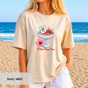 Peut inclure: Un t-shirt ivoire avec un motif repr&eacute;sentant une coupe &agrave; pied bleu clair remplie de glace rose, une tranche de past&egrave;que, une cerise et une tuile de mah-jong avec le caract&egrave;re dragon vert. Une fleur d'hibiscus rose et le texte Mahjong Happy Hour.