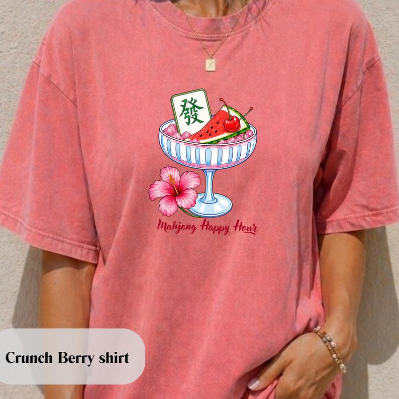 Peut inclure: Un t-shirt &agrave; manches courtes couleur corail avec un motif repr&eacute;sentant une coupe en verre bleu clair remplie de glace, une tranche de past&egrave;que, une cerise et une tuile de mahjong verte. Une fleur d'hibiscus rose et le texte Mahjong Happy Hour.