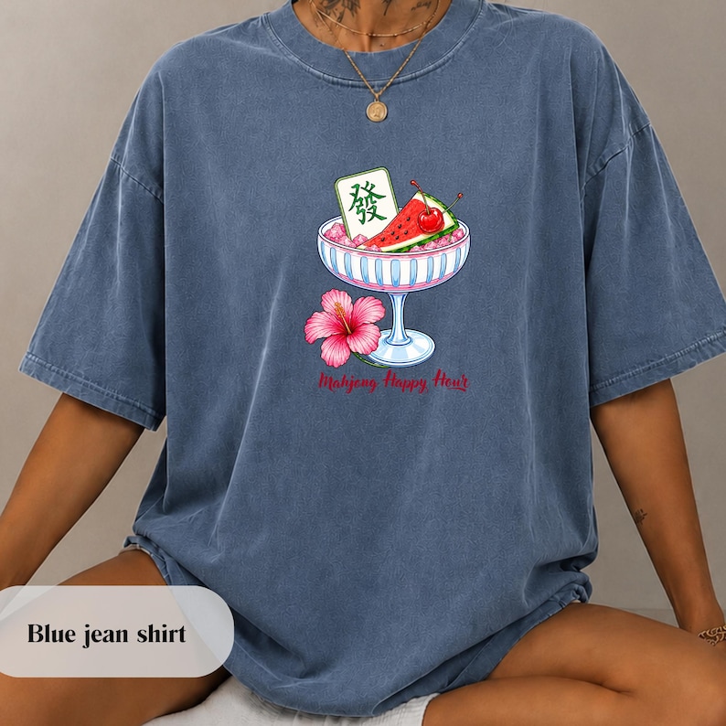 Peut inclure: Un t-shirt bleu d&eacute;lav&eacute; arborant un motif de coupe en verre contenant des tranches de past&egrave;que, une cerise et une tuile de mahjong avec le caract&egrave;re 'fa'. Sous la coupe, une fleur d'hibiscus rose et le texte 'Mahjong Happy Hour'.