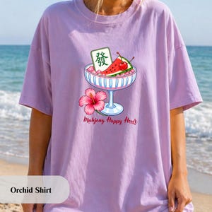 Peut inclure: Un t-shirt couleur orchid&eacute;e avec un motif repr&eacute;sentant un bol en verre rempli de glace, une tranche de past&egrave;que, une cerise rouge et une tuile de mah-jong verte avec le caract&egrave;re Fa. Une fleur d'hibiscus rose est sous le texte Mahjong Happy Hour.