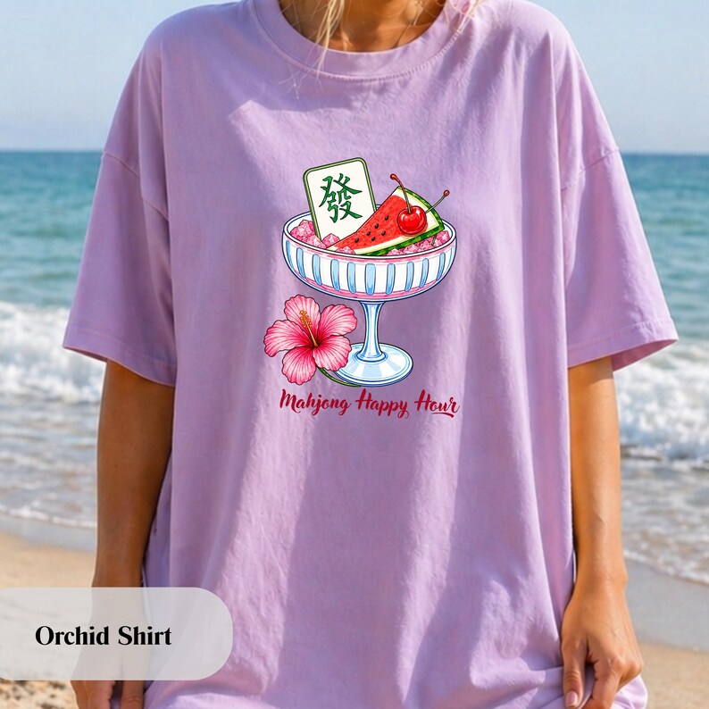 Peut inclure: Un t-shirt couleur orchid&eacute;e avec un motif repr&eacute;sentant un bol en verre rempli de glace, une tranche de past&egrave;que, une cerise rouge et une tuile de mah-jong verte avec le caract&egrave;re Fa. Une fleur d'hibiscus rose est sous le texte Mahjong Happy Hour.