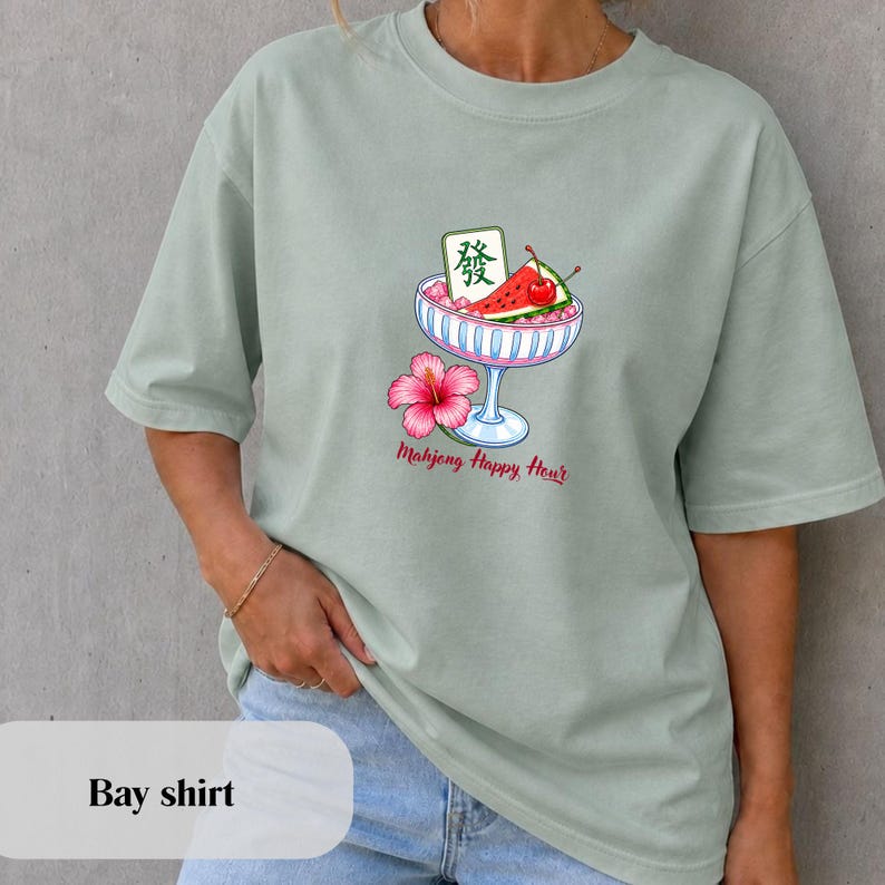 Peut inclure: Un t-shirt vert sauge avec un motif repr&eacute;sentant un bol en verre rempli de glace pil&eacute;e rose, une tranche de past&egrave;que, une cerise et une tuile de mah-jong verte avec le caract&egrave;re Fa. Une fleur d'hibiscus rose et le texte Mahjong Happy Hour.