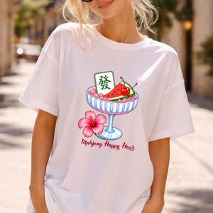 Peut inclure: Un t-shirt blanc avec un motif repr&eacute;sentant un bol en verre &agrave; bord bleu rempli de glace rose, une tranche de past&egrave;que, une cerise rouge et une tuile de mah-jong verte avec le caract&egrave;re "發". Une fleur d'hibiscus rose et le texte "Mahjong Happy Hour".