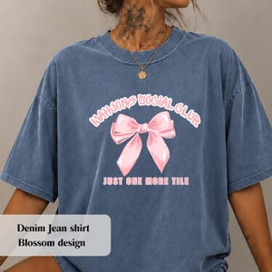 May include: A blue denim-coloured t-shirt featuring a graphic print of a light pink bow in the centre. Above the bow, the text reads "MAHJONG SOCIAL CLUB" in a curved white font, and below the bow, the text reads "JUST ONE MORE TILE" in white.